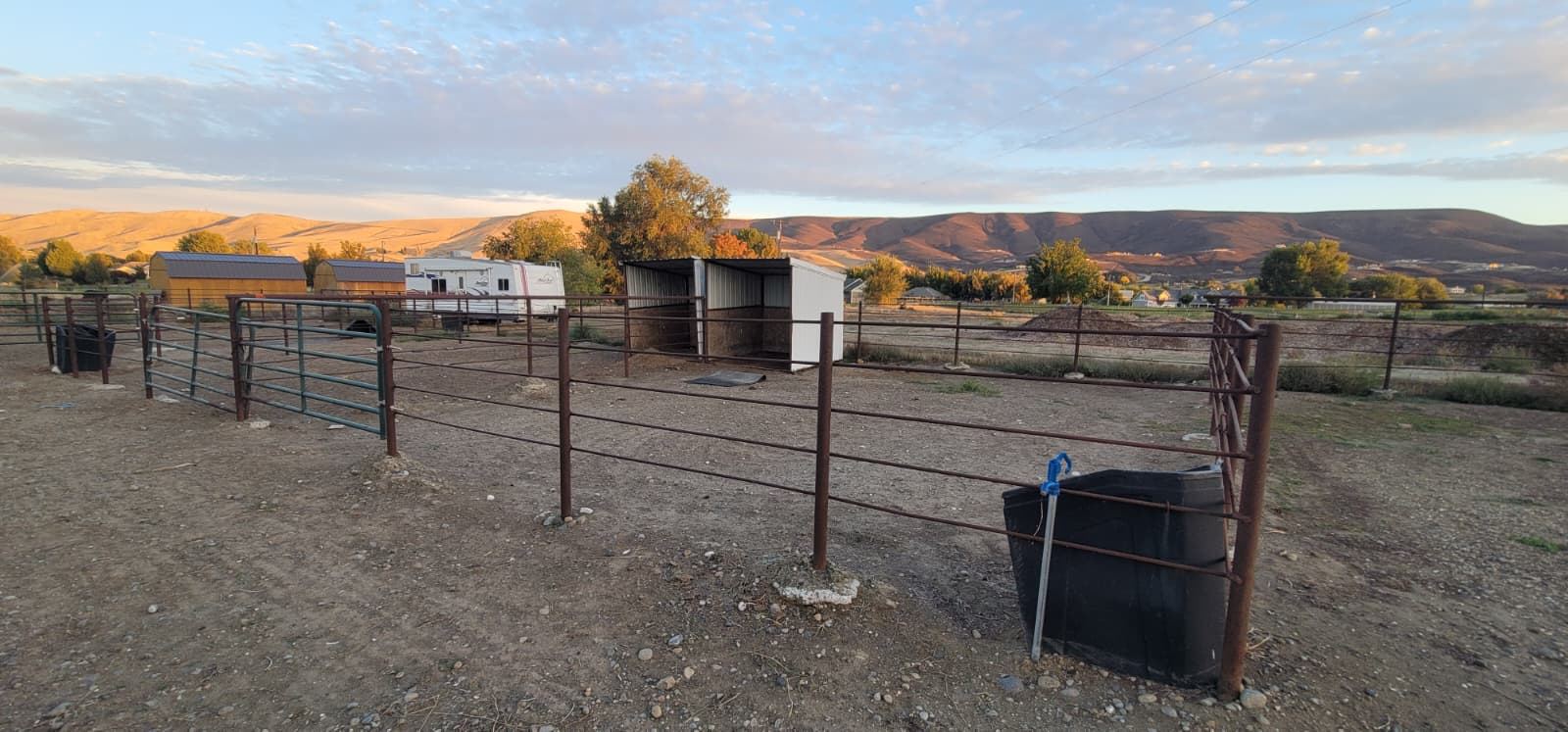 Pipe-fenced paddock with a black water tub, a white run-in shelter, an RV and sheds behind, and brown hills under an evening sky