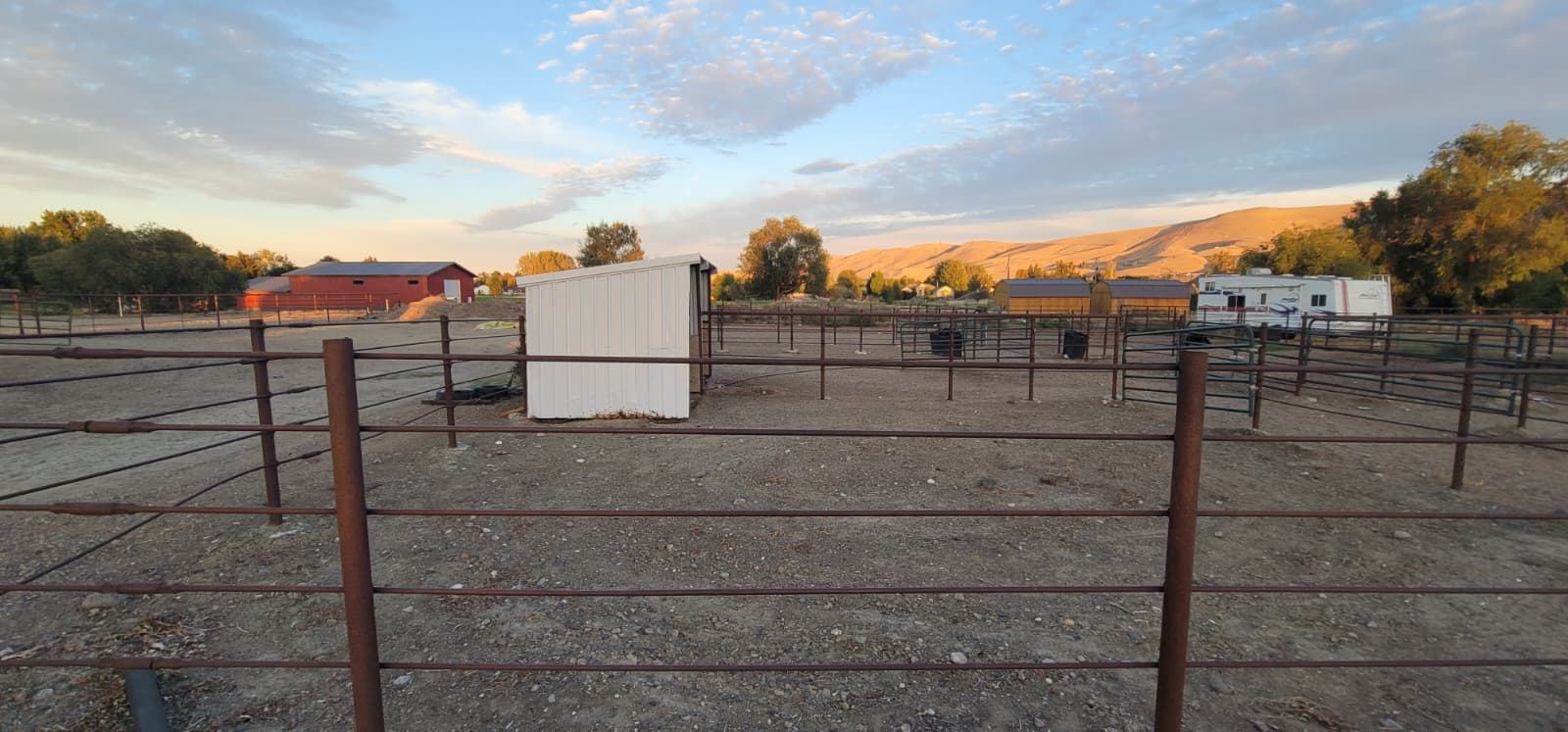 Several pipe-fenced dirt paddocks with a white shelter, a red barn and an RV, with hills behind