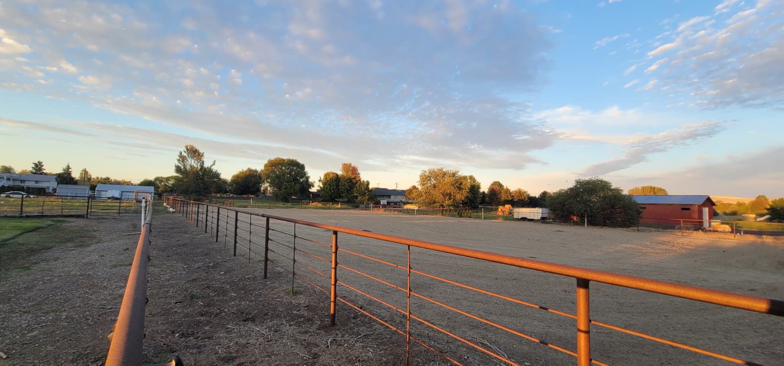 Long sand arena with orange pipe fencing at golden hour, trees and a red barn in the distance