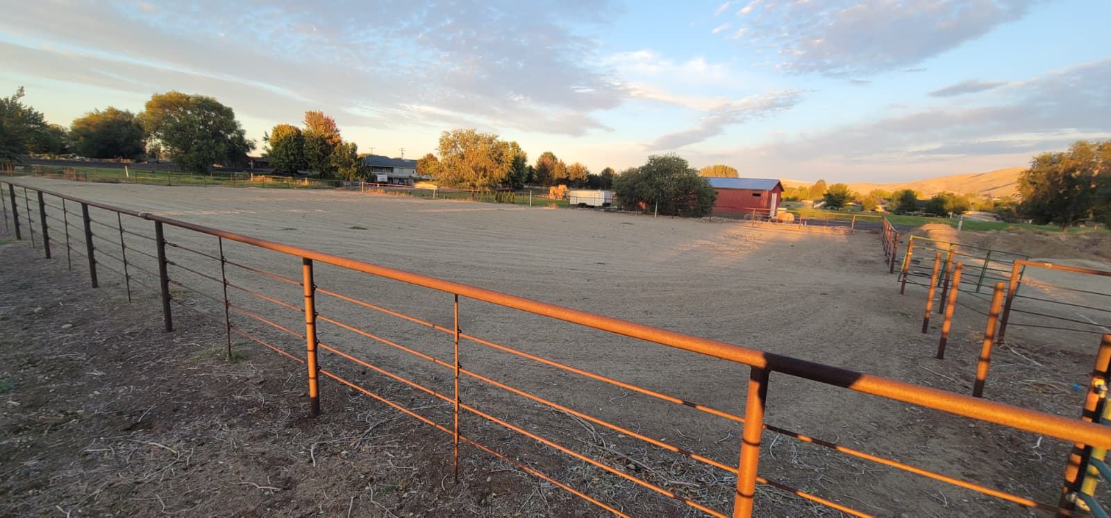 Sand arena seen from the corner of its orange pipe fence, with a red barn, a trailer and trees beyond